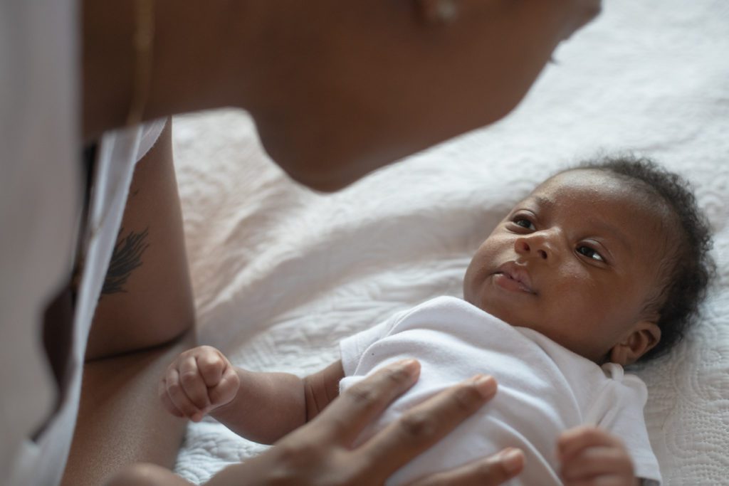 An infant African boy lays on a bed looking up at his mother.  The young mother is leaning in close to his face and is resting her hand on his tummy as she looks down at him tenderly.  They are both dressed casually.